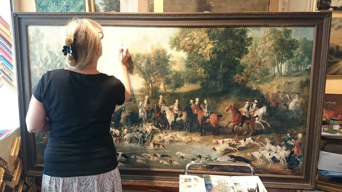 Photo où Véronique fait des retouches picturales sur un grand tableau de coloré, rempli de personnages à cheval et d'une grande quantité de chiens de chasse